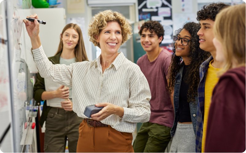 Female teacher writing on a whiteboard surrounded by teenage students
